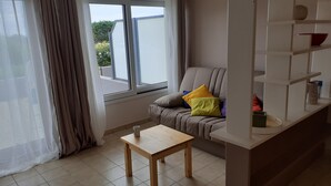 A living room with a grey sofa, a wooden table, and a window with curtains. - Studio Vue mer RDC à 100 m de la Plage (Larmor-Plage)