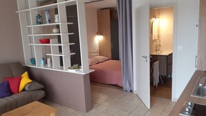 A compact living space with a kitchenette, a bed with a pink bedspread, and a sofa with colorful cushions. - Studio Vue mer RDC à 100 m de la Plage (Larmor-Plage)