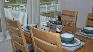 Mesa de comedor lista para cuatro personas con sillas de madera, una botella de vino y vista a la playa a través de ventanas amplias.