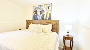 Un lit bien fait, avec un couvre-lit blanc et des oreillers, une tête de lit en bois, une table de chevet avec une lampe et un tableau au-dessus du lit.