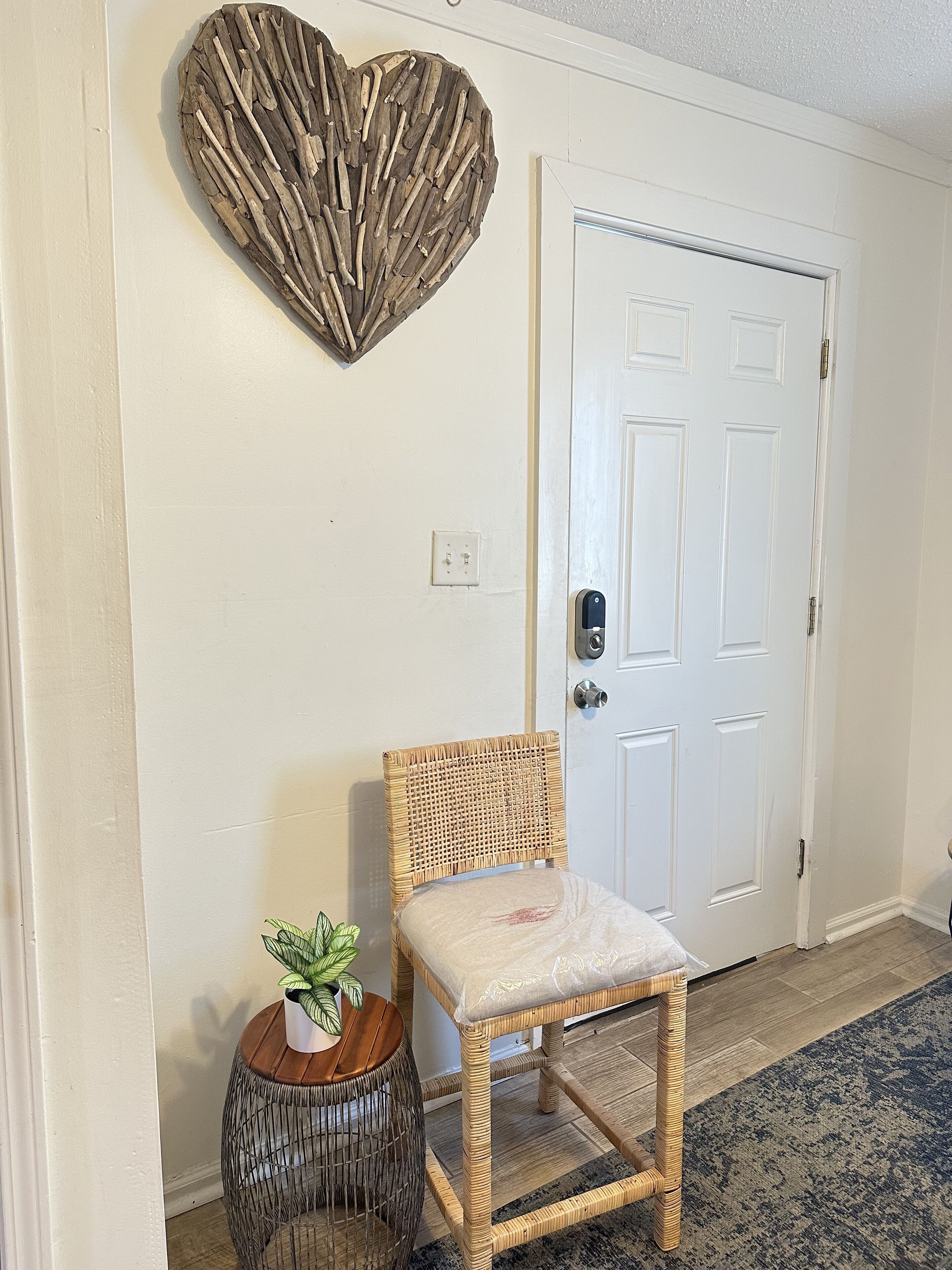 A room with a wicker chair, a small wooden side table with a plant, and a decorative heart-shaped wall art above a white door.
