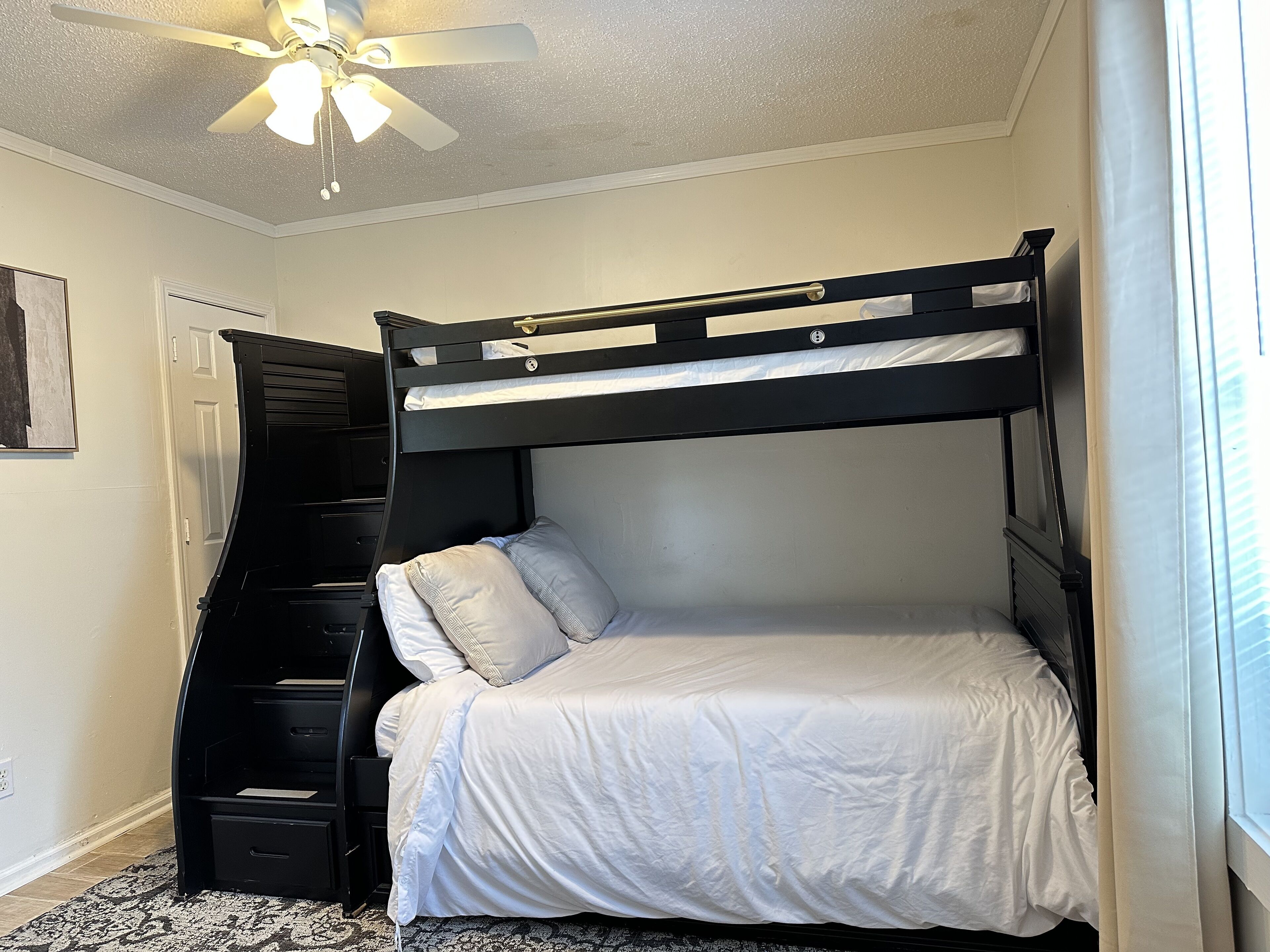 A bunk bed with a desk and drawers, a ceiling fan, and a window with curtains.