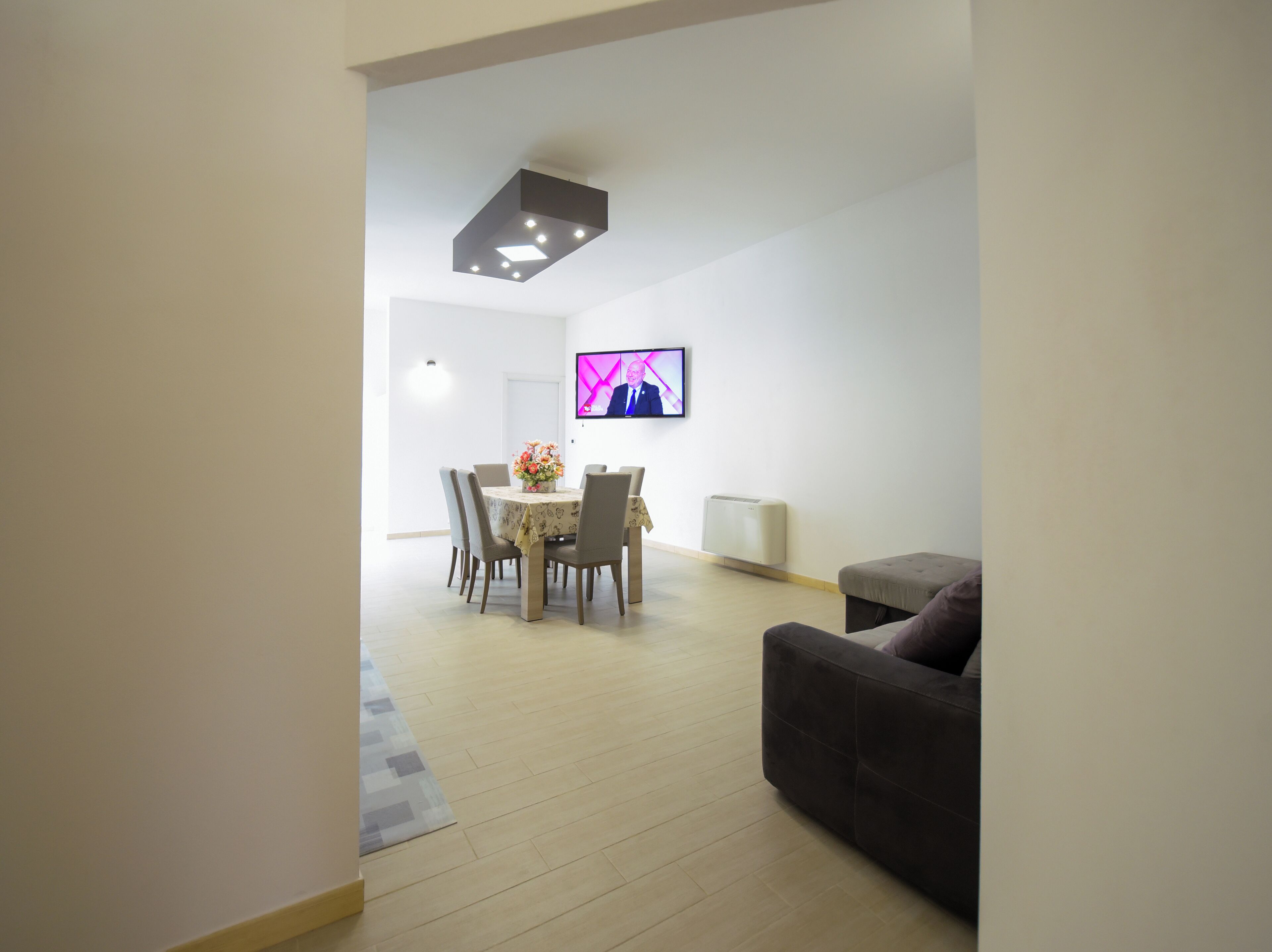 A modern living room with a dark sofa, a dining area with a table and chairs, and a flat-screen TV mounted on the wall.