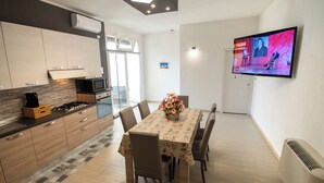 A modern kitchen with wooden cabinets, a gas stove, and a microwave. There is a dining table with chairs and a floral tablecloth. A flat-screen TV is mounted on the wall. - Casa A&F - Fra (Torricella)
