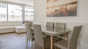 A dining area with a glass table and wicker chairs, a white sofa, and a large window with a view of buildings and a sunset.