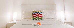 Een net opgemaakt bed met witte lakens, een kleurrijk zigzagkussen en een nachtkastje met een lamp.