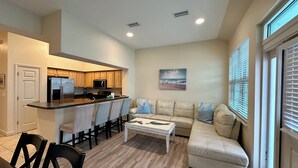 A modern living room with a beige sofa, a coffee table, and a kitchen area with wooden cabinets and stainless steel appliances. - Beach Retreat in Perdido Key, Florida (Pensacola)