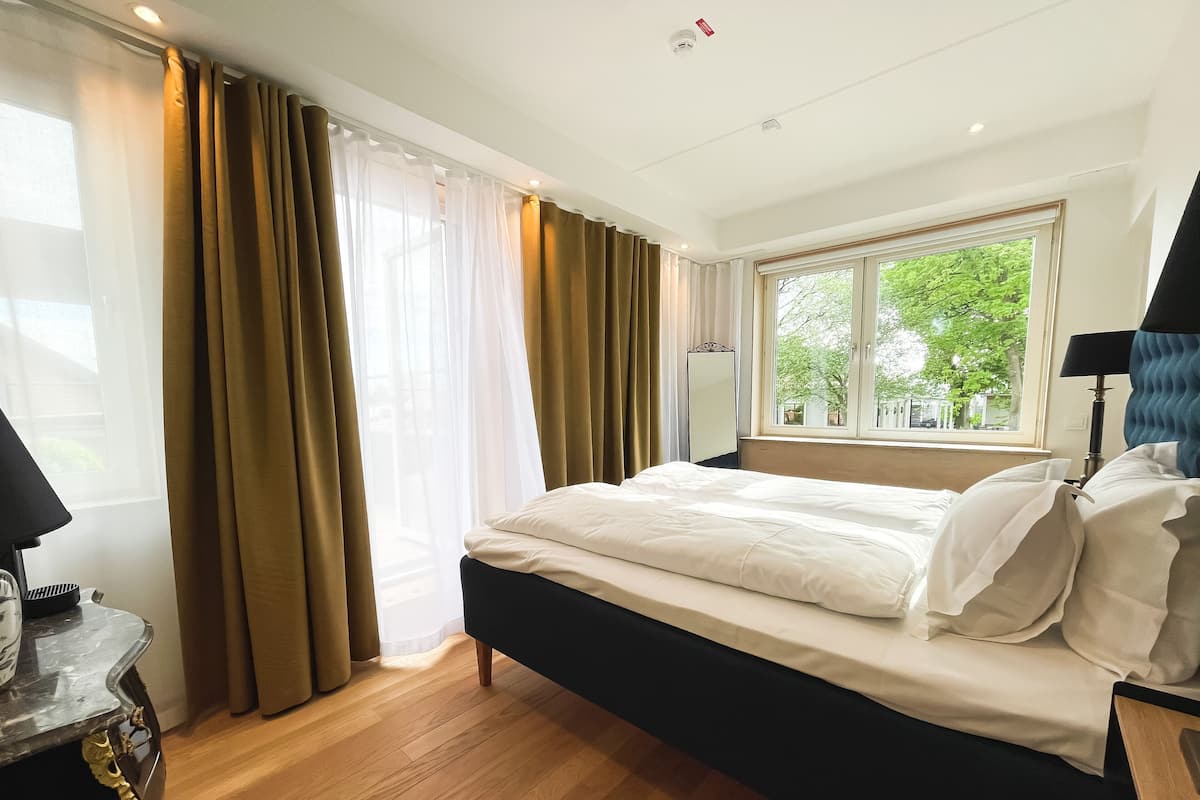 A hotel room with a bed, a window with a view of greenery, a bedside table with a lamp, and a mirror.