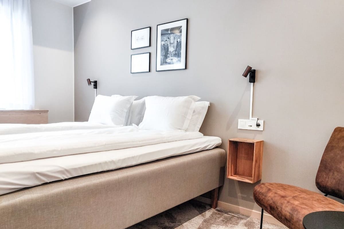 A modern hotel room with a bed, a wooden nightstand, a chair, and a wall-mounted lamp.