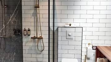 Un baño moderno con ducha envuelta en vidrio, paredes de azulejos blancos, suelo de mármol y tocador de madera con inodoro.