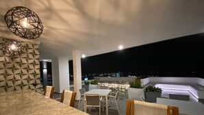 Een moderne eethoek buiten met een bar, zitplaatsen en uitzicht over de stad 's nachts.