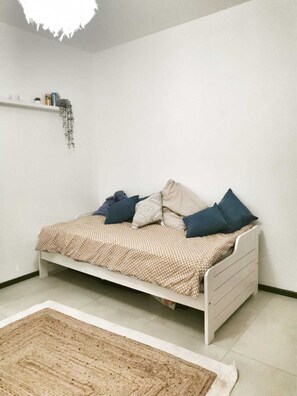 Quarto individual com uma cama branca, roupa de cama bege e estampada, e um tapete pequeno.