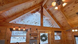 Interno di una baita in legno con soffitto alto, travi a vista e un lucernario. Si vedono finestre con vetro satinato, un camino e un angolo cottura con lavello e piano di lavoro.