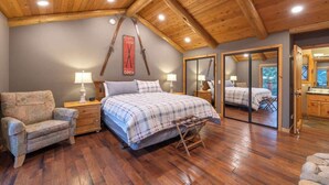 Une chambre avec un plafond en bois, un lit recouvert d’une couverture à carreaux, une chaise, une table de chevet avec une lampe et un miroir.