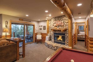 L’intérieur chaleureux d’un chalet en bois, avec une cheminée en pierre, une table de billard et un coin repas offrant une vue sur l’extérieur.