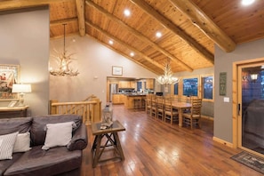 A spacious living area with a wooden ceiling, a dining table with chairs, a kitchen in the background, and a large window.
