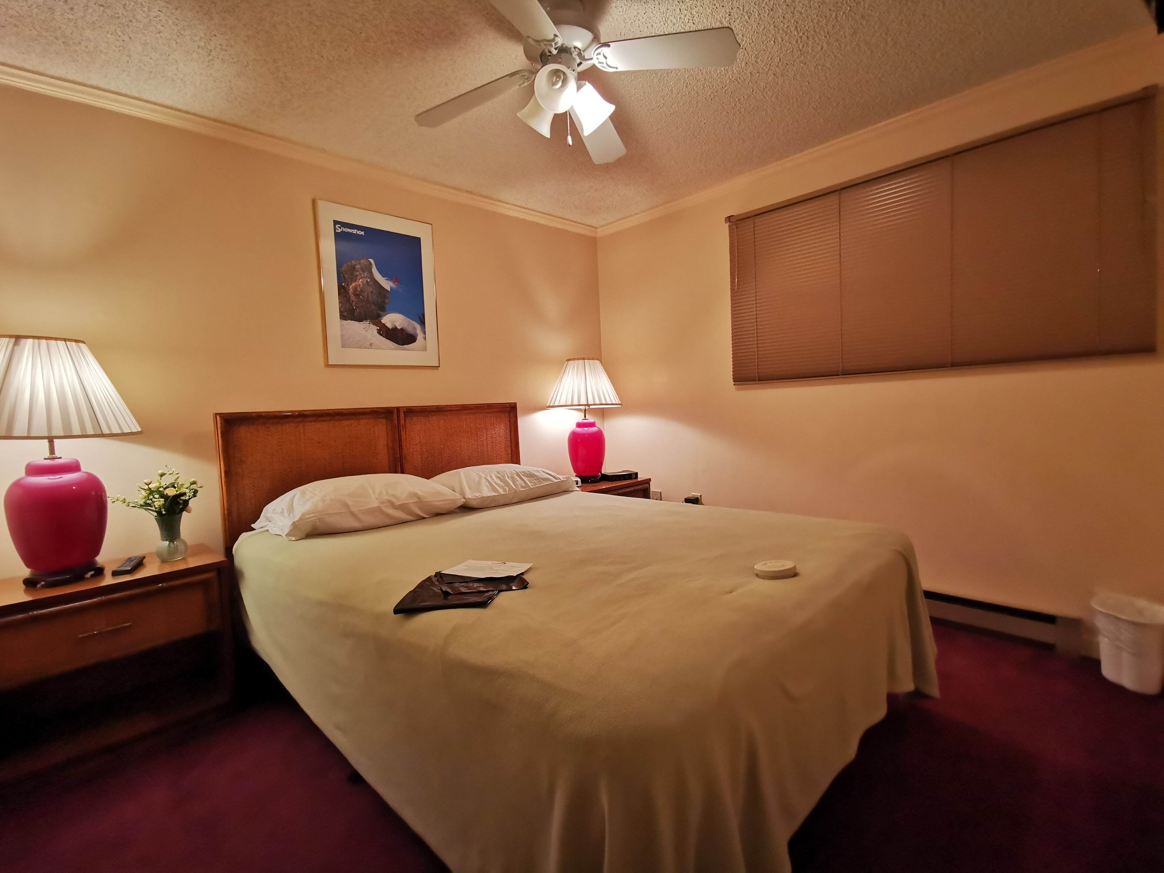 A hotel room with a bed, two bedside lamps, a ceiling fan, a framed picture on the wall, and a window with blinds.