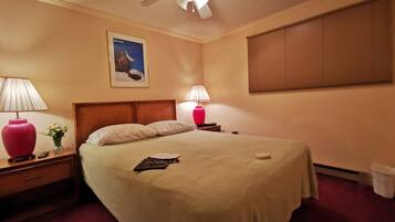 A hotel room with a bed, two bedside lamps, a ceiling fan, a framed picture on the wall, and a window with blinds.