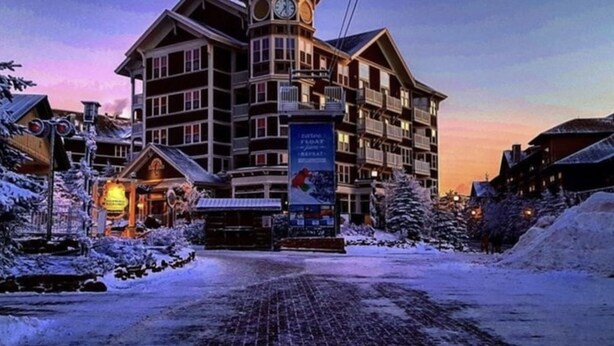 Een hotel met meerdere verdiepingen, een opvallende klokkentoren, een reclame voor een skilift en een besneeuwd landschap.
