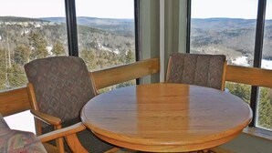 Een ronde houten tafel met vier stoelen, grote ramen met uitzicht op een bebost landschap.