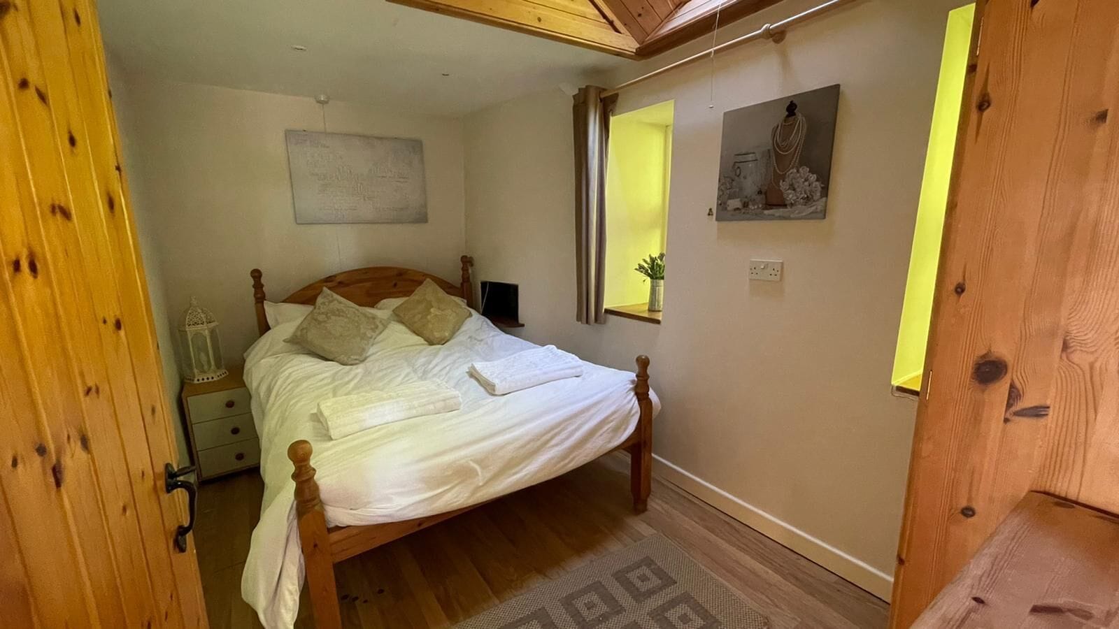 Une chambre à coucher avec un lit en bois, une table de chevet, une commode, une fenêtre avec des rideaux et une œuvre d’art accrochée au mur.