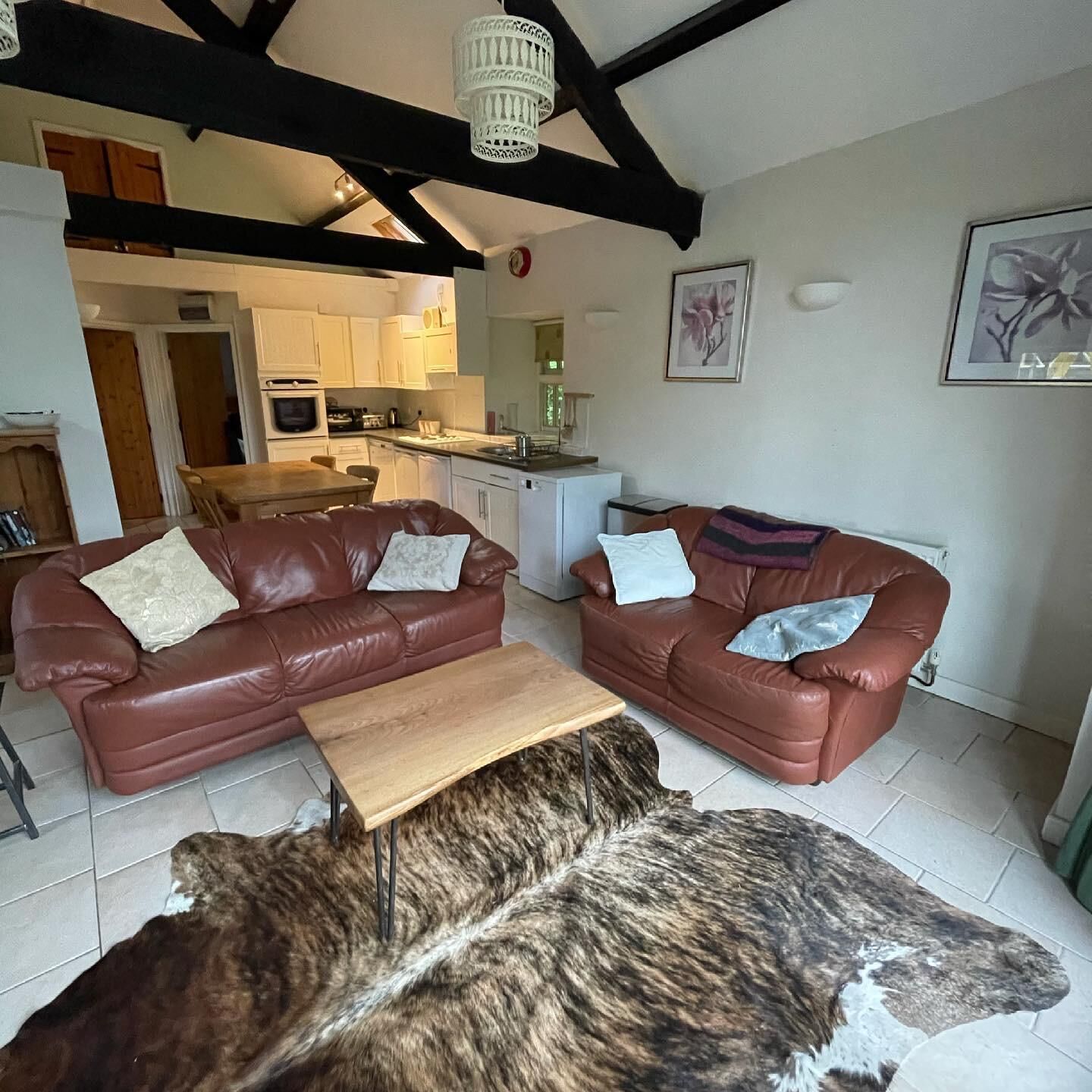 A living room with two leather sofas, a wooden coffee table, and a fur rug. There is a kitchen in the background with white cabinets and a white refrigerator.