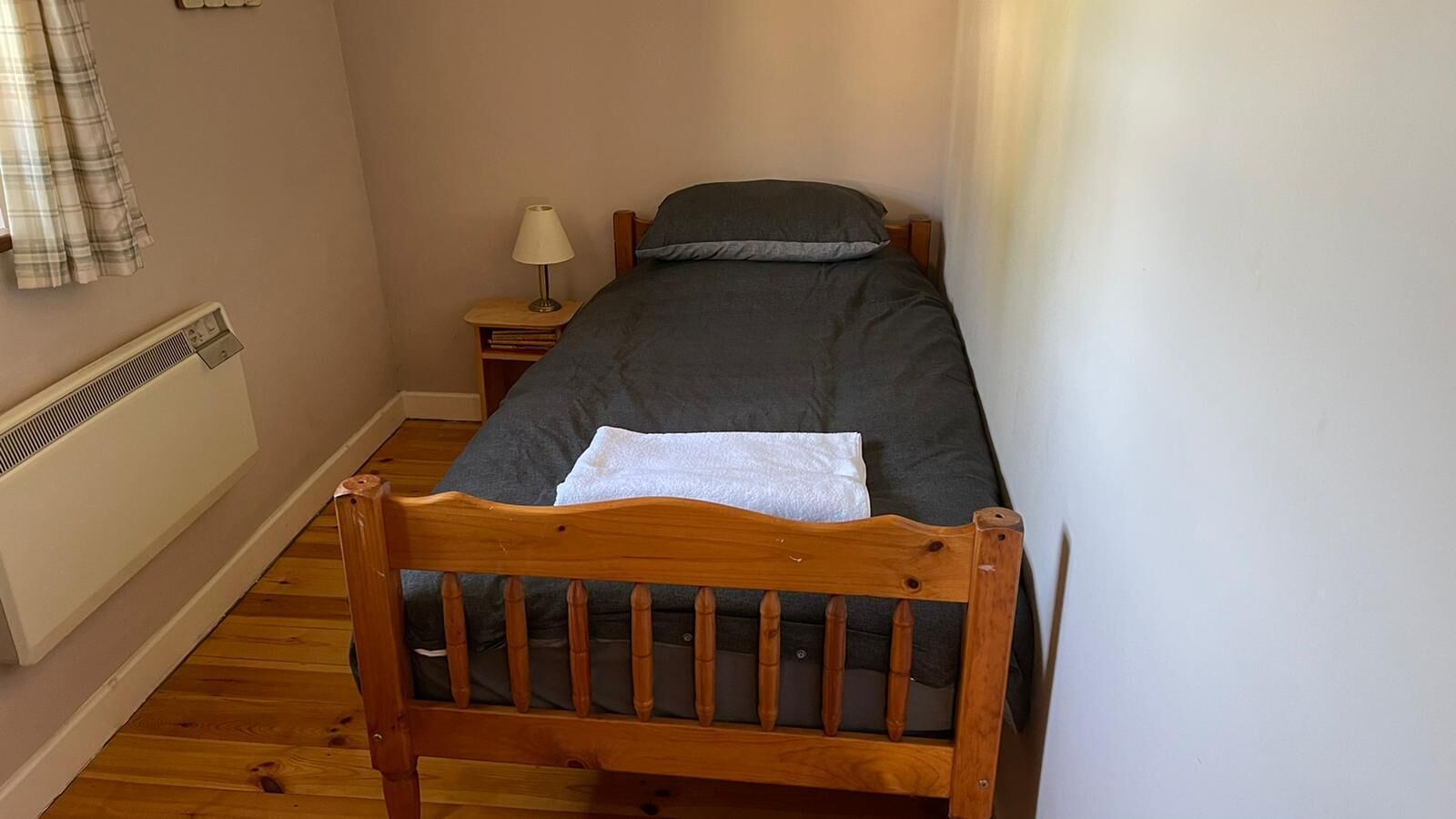 A small, single-bed room with a wooden headboard, a bedside table with a lamp, and a radiator on the wall.