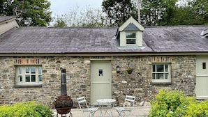 Une petite maison en pierre avec une cheminée, une petite fenêtre et une porte. Il y a une table et des chaises à l’extérieur.