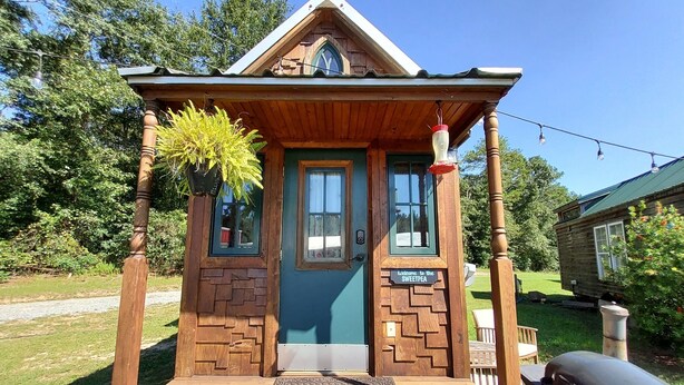Une petite construction en bois avec une porte verte, une pancarte indiquant «Bienvenue» et un porche doté d’un banc.