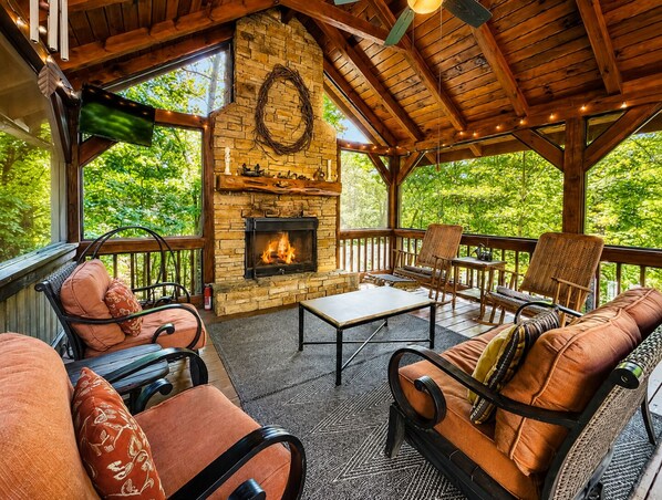 A cozy outdoor seating area with a stone fireplace, wooden furniture, and a view of greenery. - Tumnus Ridge - Super Dog Friendly | Hot tub | Fire Pit (Morganton)