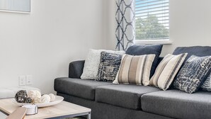 A modern living room with a grey sofa, patterned cushions, a white side table with decorative items, and a window with blinds. - Downtown Modern Bungalow (Dallas)