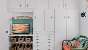 A room with a TV on a stand, white cabinets with drawers, wicker baskets, and a green chair with a cushion.