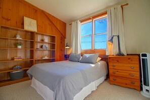 Ein Schlafzimmer mit einem Bett, einem Schminktisch, einem Nachttisch, einer Lampe, einem Fenster mit Vorhängen und einer Holzwandregal mit Büchern und Dekoration.