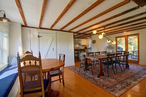 A dining area with a wooden table and chairs, a kitchen in the background, and a view of the outdoors through a glass door. - Renovated Farmhouse < 1 Mile to Okemo Mountain! (Ludlow)