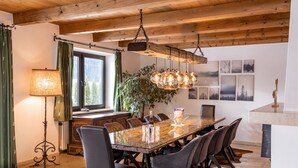 A dining area with a wooden table, chairs, a chandelier, and a large window with curtains. - Exclusive Villa Tirol (Fieberbrunn)