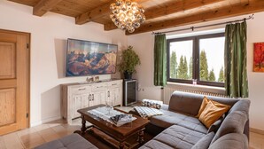 A living room with a wooden ceiling, a large flat-screen TV, a sofa, a coffee table, and a window with curtains. - Exclusive Villa Tirol (Fieberbrunn)