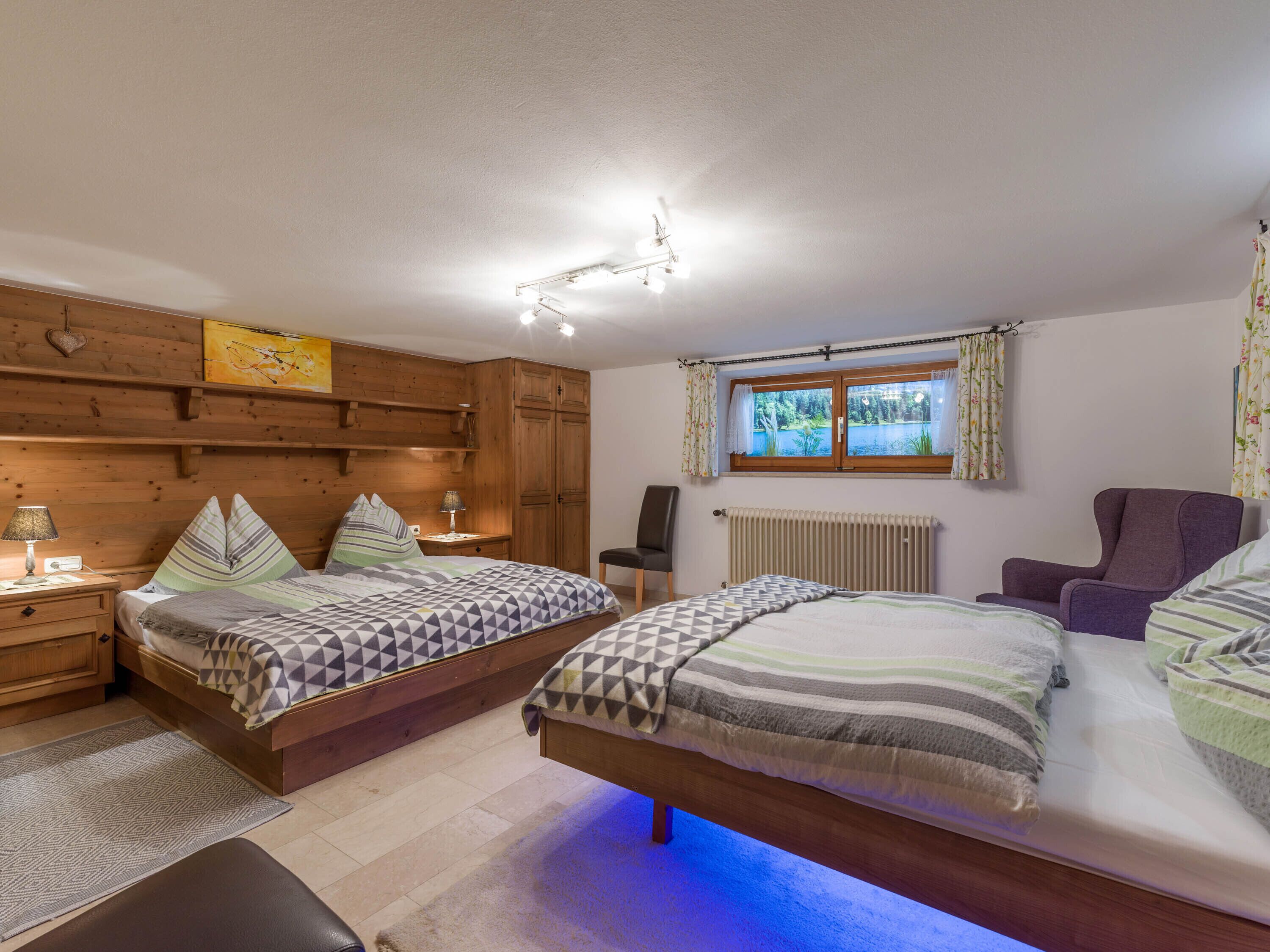 Quarto com duas camas, cabeceira de madeira, janela com cortinas, radiador e uma cadeira.