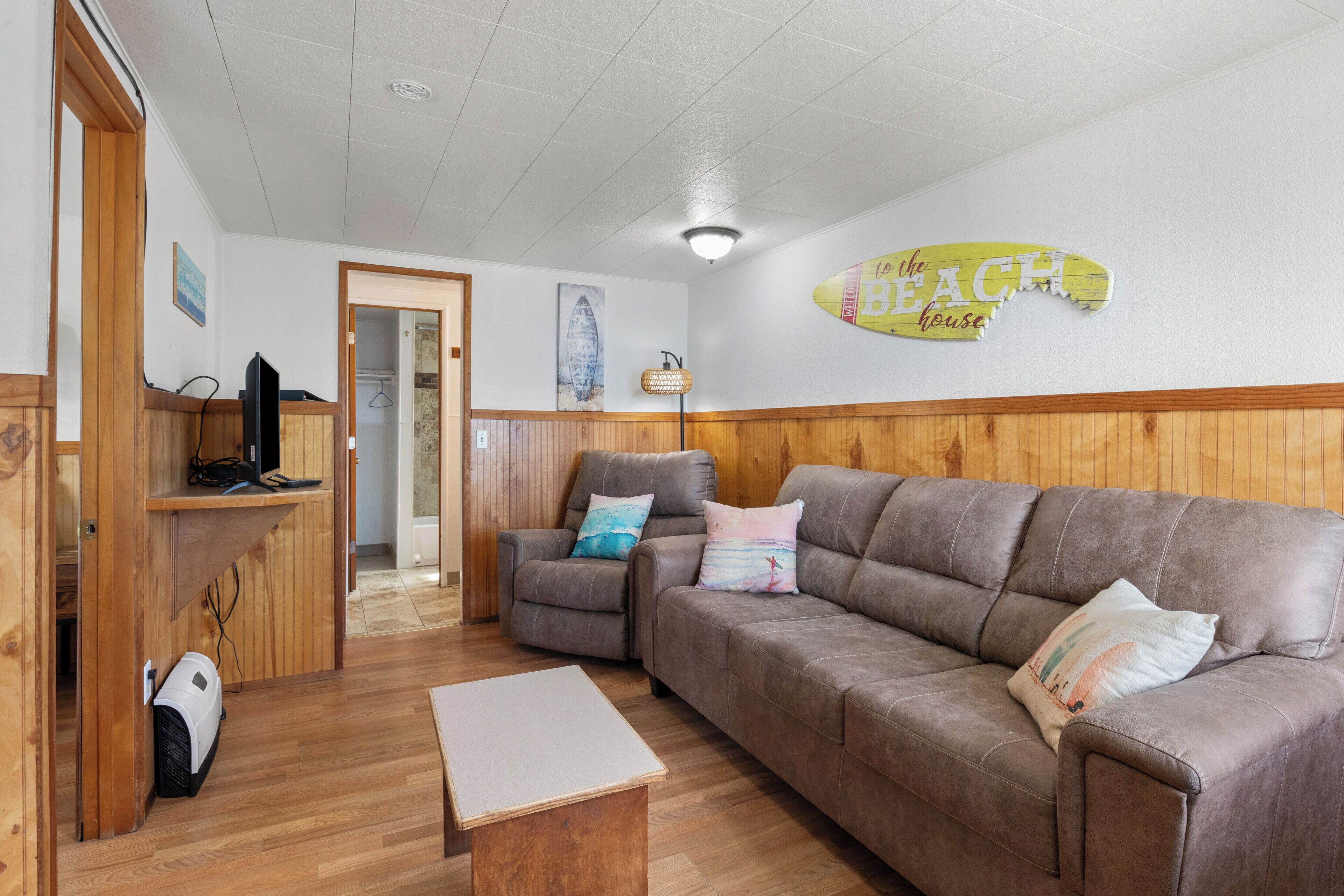 Une pièce avec un canapé en cuir marron, une table basse en bois, un téléviseur posé sur un meuble en bois, et une pancarte murale indiquant « vers la maison de plage ».