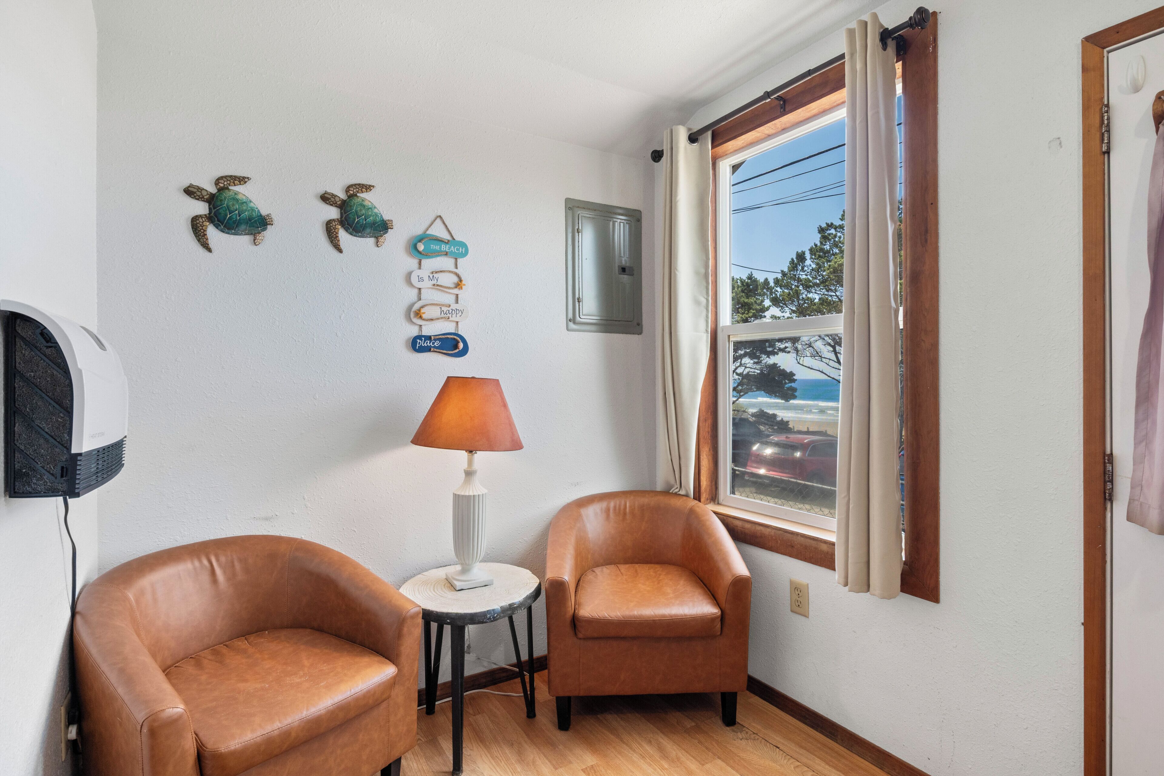 A room with two leather chairs, a small table with a lamp, decorative wall art, a window with curtains, and a wall-mounted air conditioner.