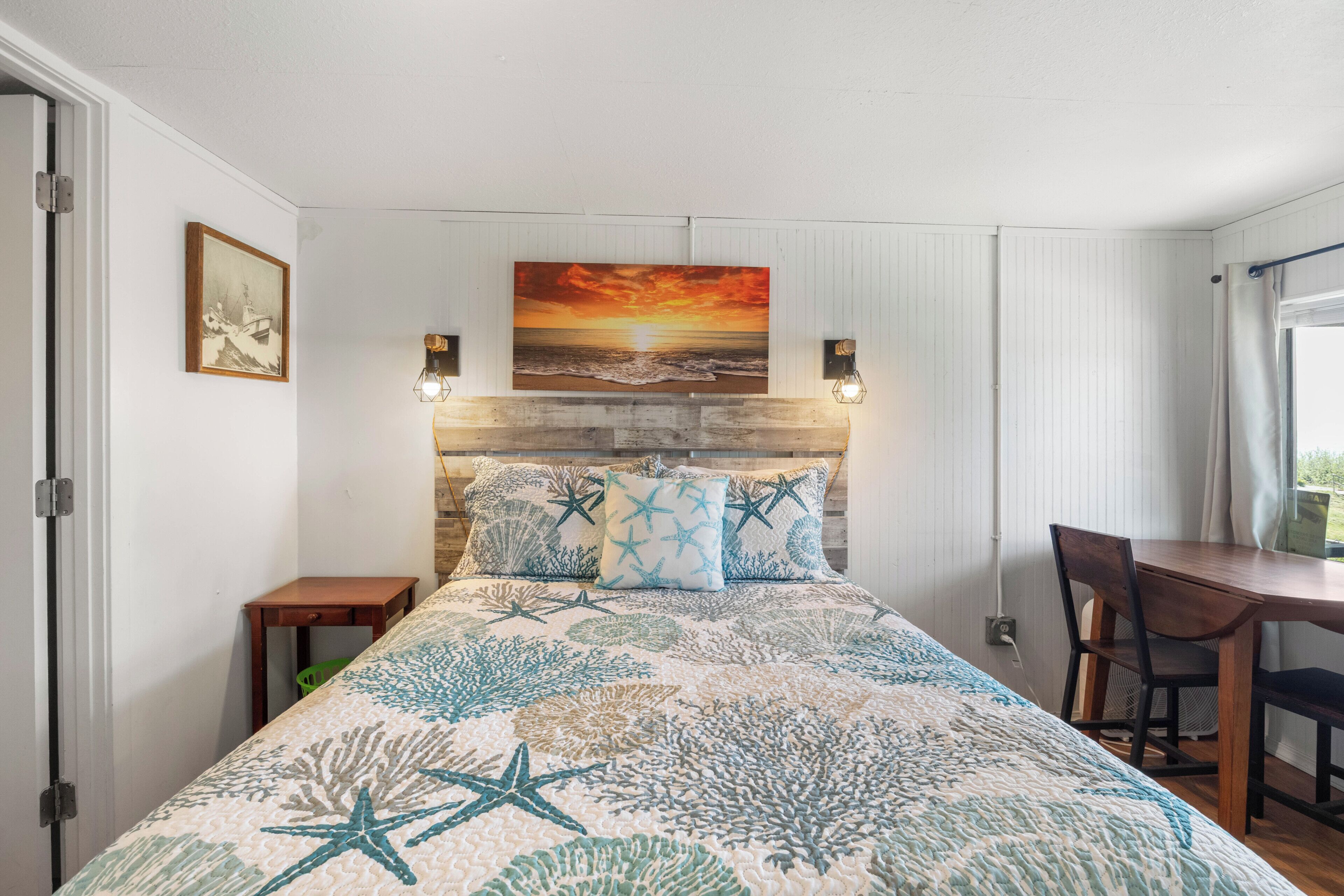 Une chambre à coucher avec un lit, un tableau représentant un coucher de soleil, un bureau et une chaise.