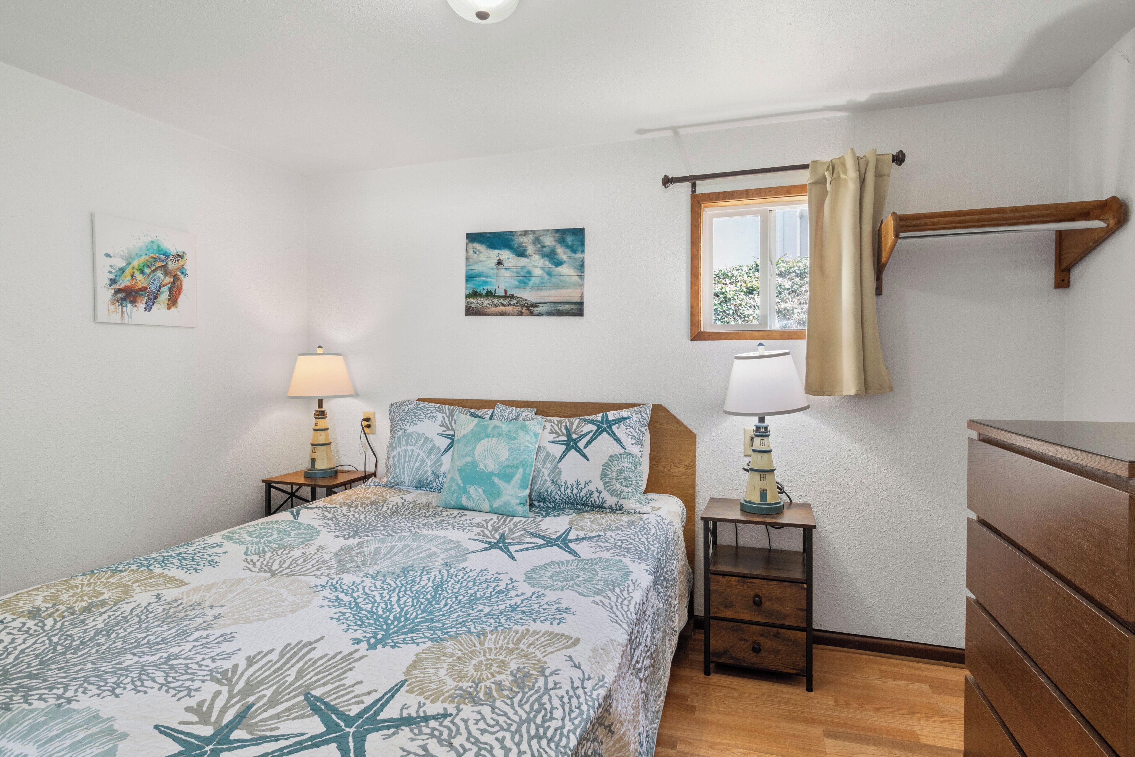A bedroom with a bed, two bedside tables with lamps, a dresser, a window with curtains, and two wall-mounted paintings.