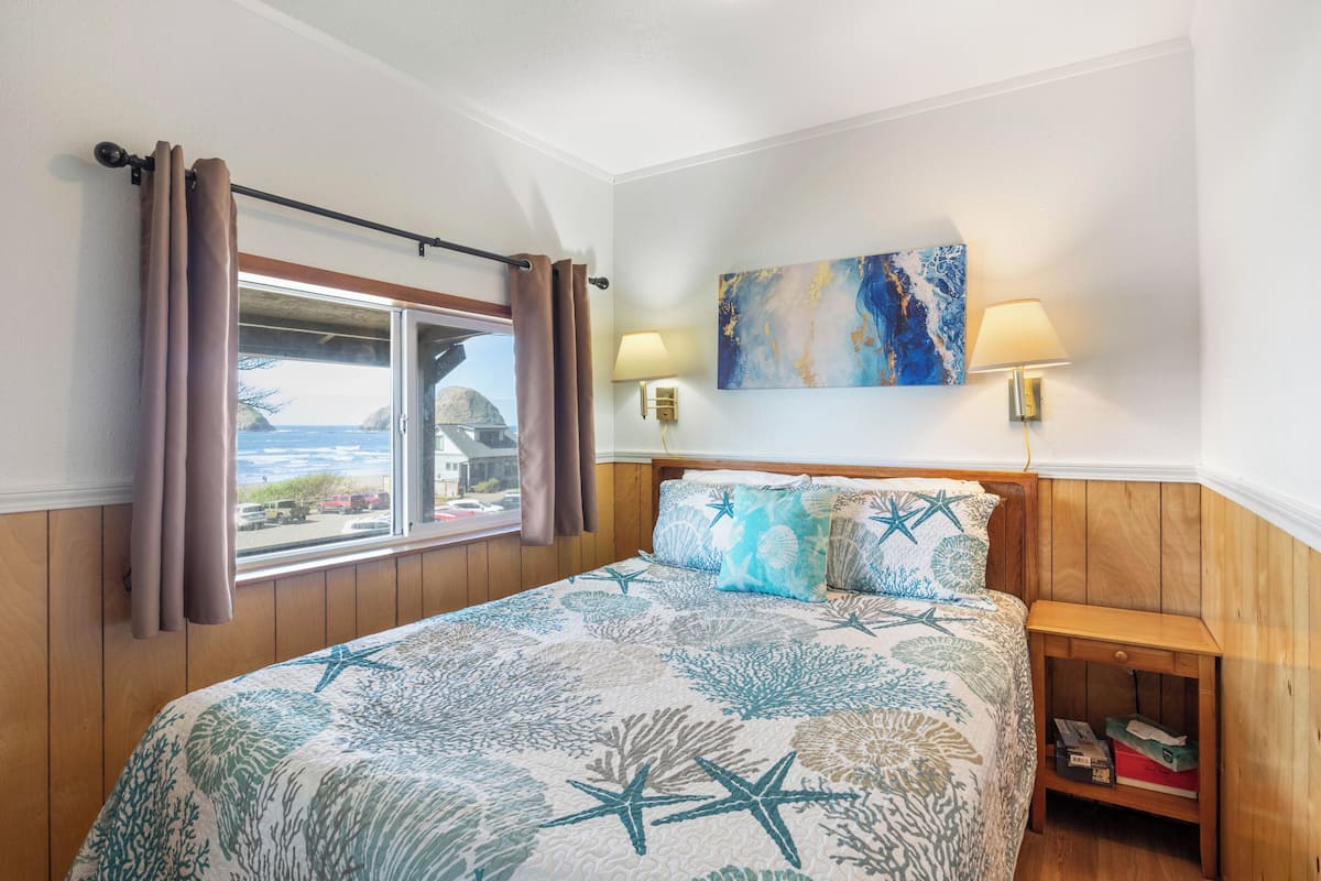 A bedroom with a bed, curtains, a window with a view, a painting, and a nightstand.