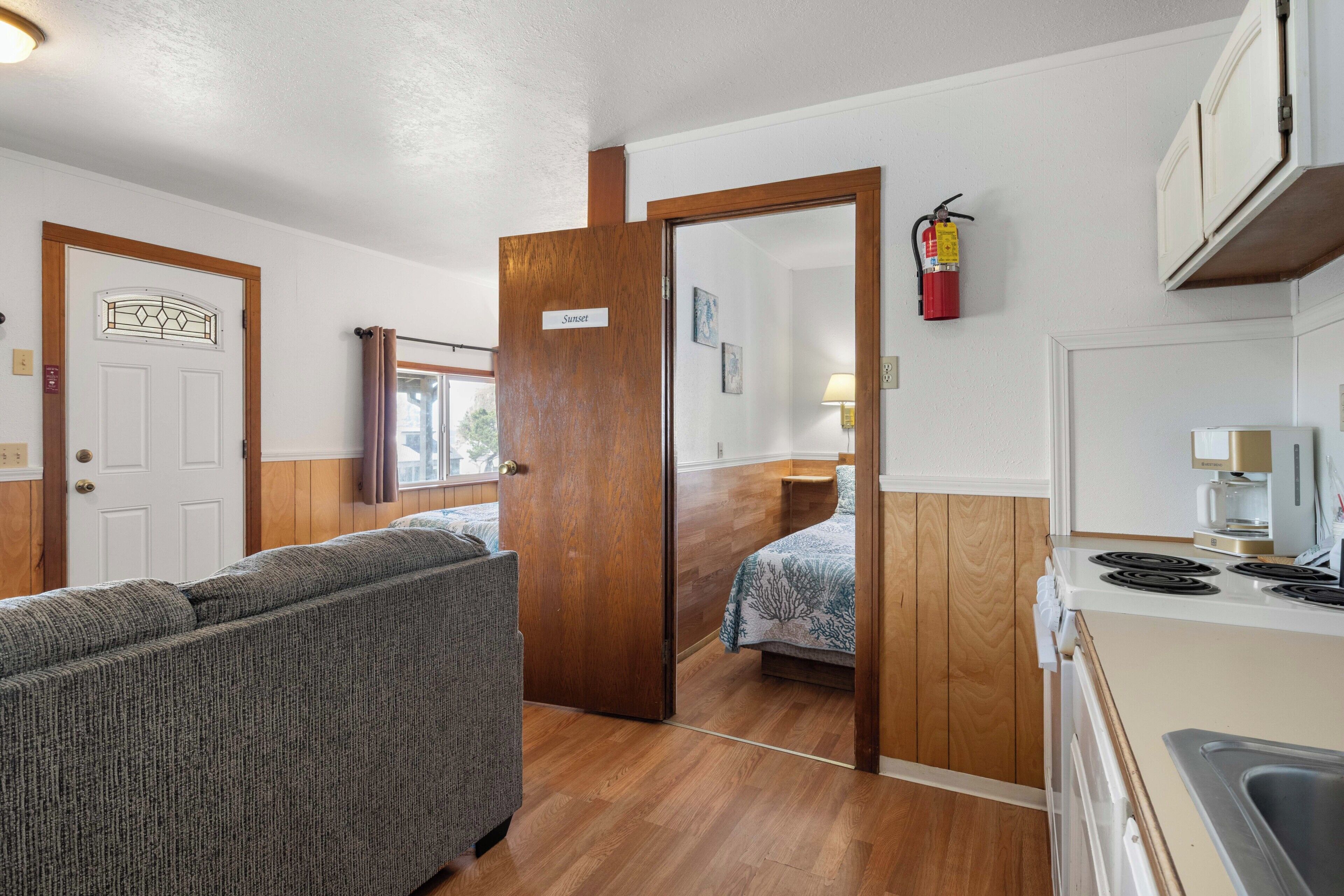A room with a sofa, a bed, a kitchen area with a stove, and a door labeled 'Sunrise.'
