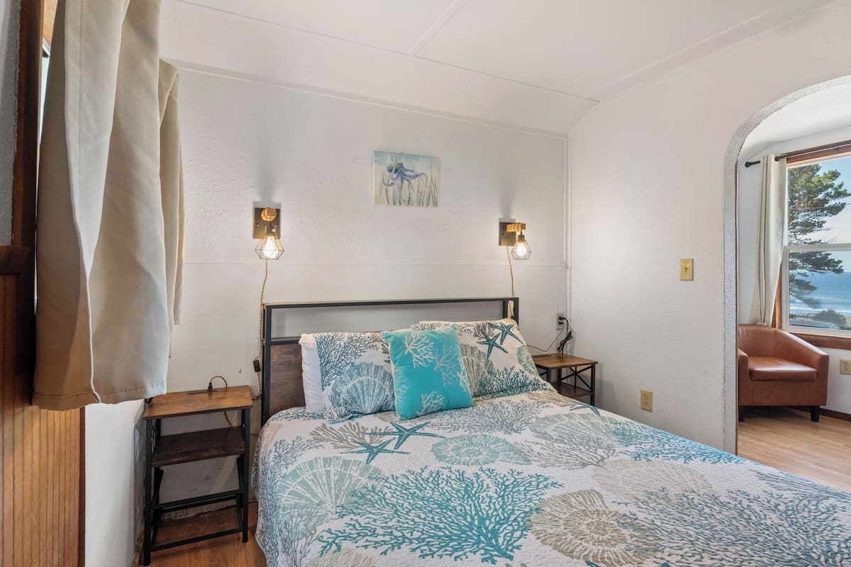 A bedroom with a bed, two wall-mounted lights, a nightstand, a chair, and a view of the outdoors.