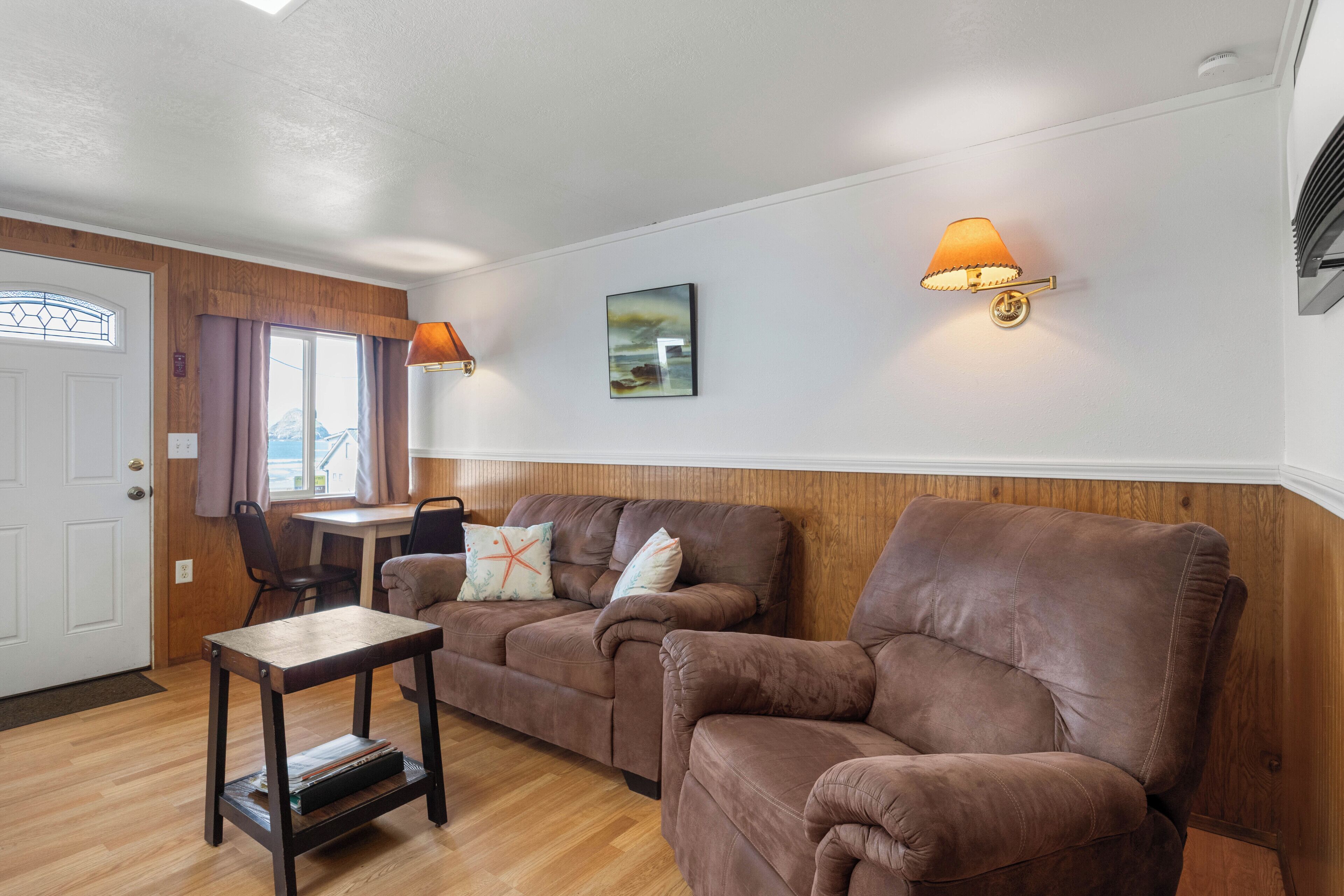 A living room with a brown sofa, a small table, a lamp, and a painting on the wall.
