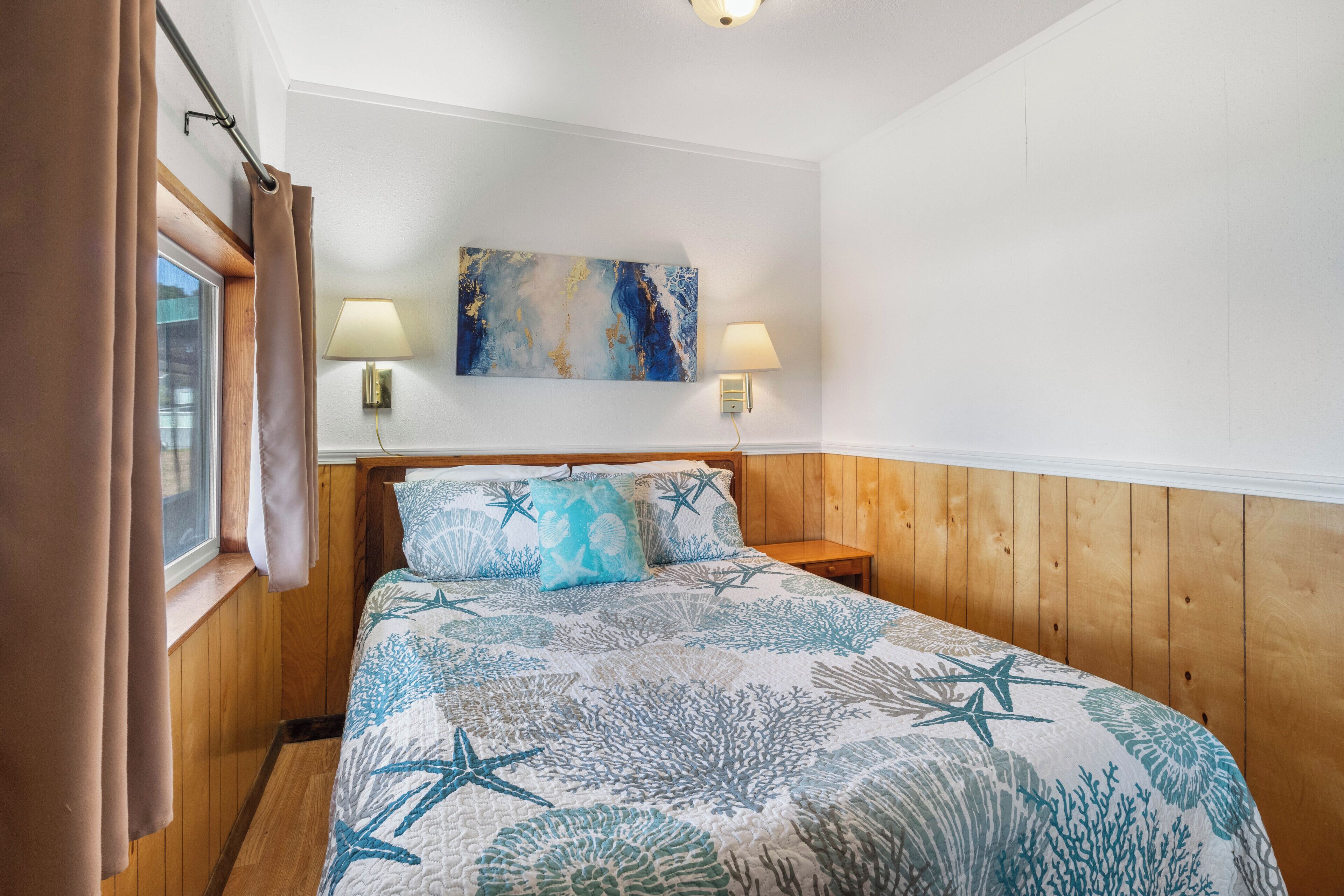 Une chambre à coucher avec une tête de lit en bois, un lit recouvert d’une couvre-lit à motifs coralliens bleus et blancs, deux lampes de chevet et un tableau accroché au mur.