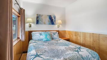 A bedroom with a wooden headboard, a bed with a blue and white coral pattern bedspread, two bedside lamps, and a painting on the wall.