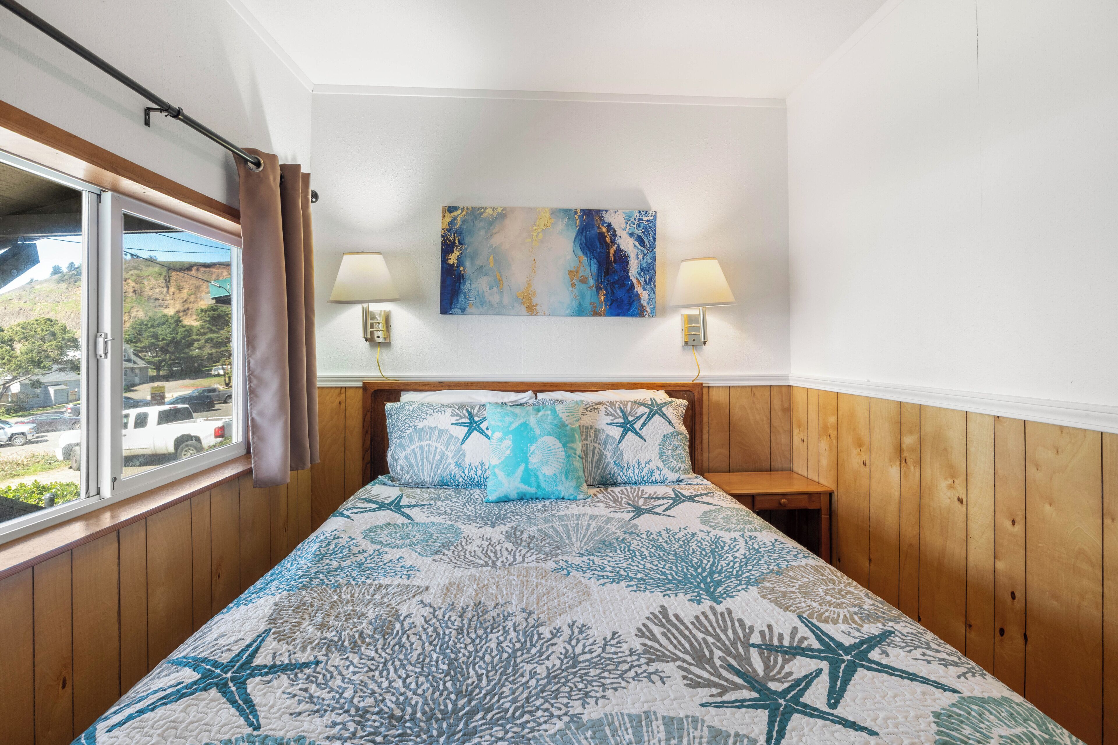 Une chambre à coucher avec un lit, deux lampes de chevet, une fenêtre avec des rideaux et un tableau accroché au mur.