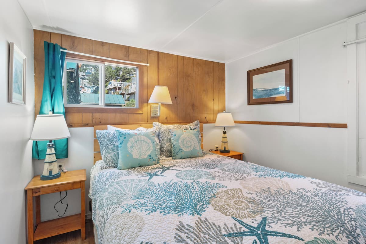 A bedroom with a bed, two bedside tables, a window with curtains, a wall-mounted lamp, and a framed picture.