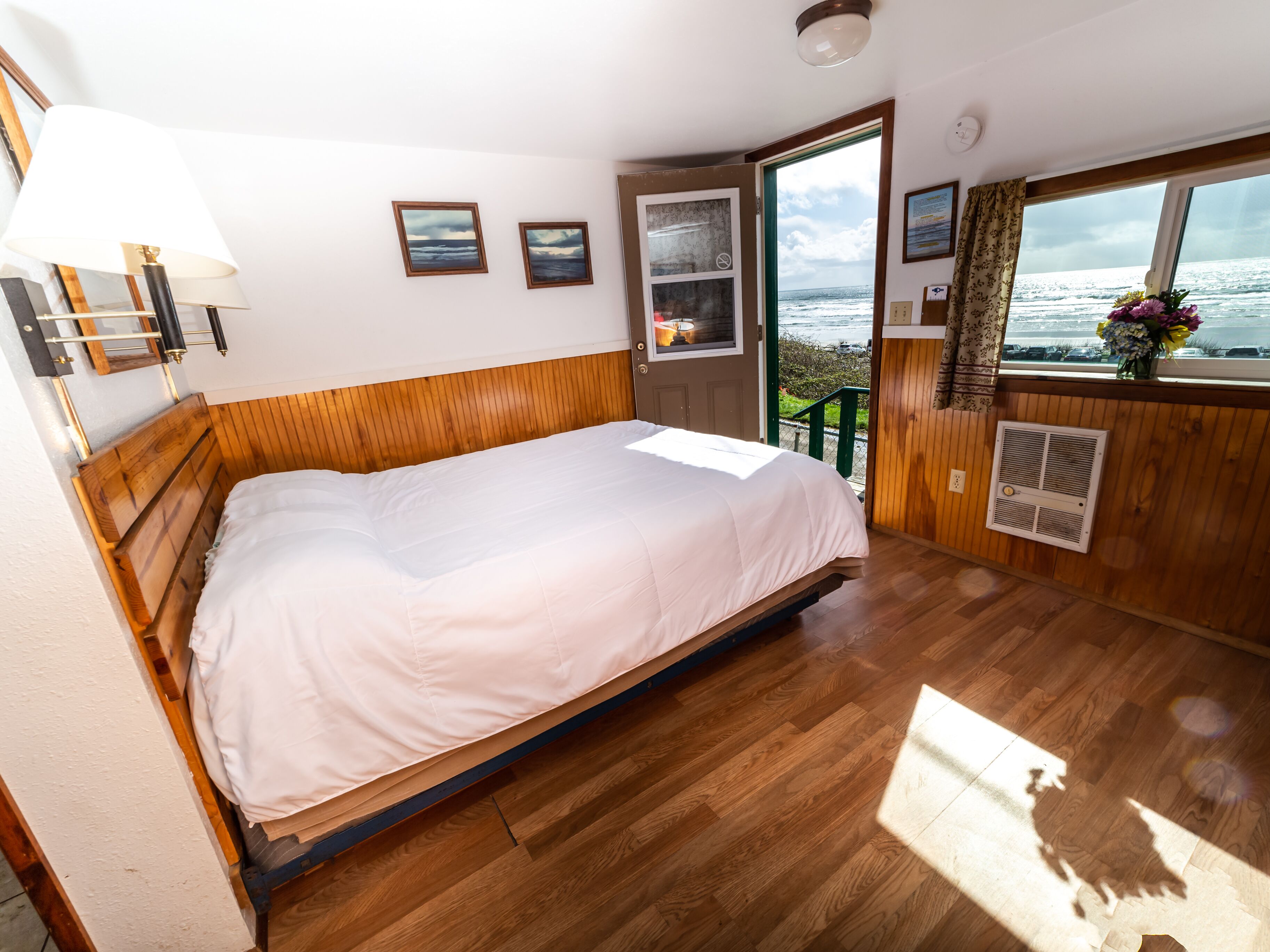 A bedroom with a wooden bed, a window with a view of the sea, and a door leading outside.